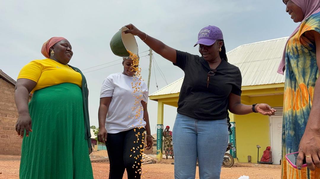 Maize grain being poured from a bowl to winnow it, watched by members of the community.