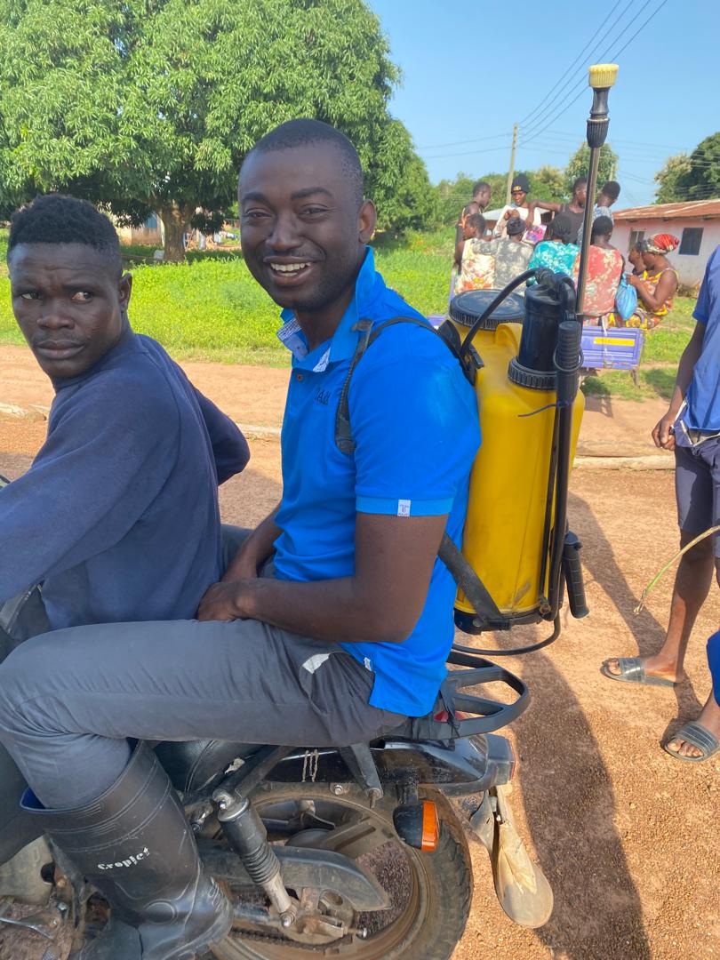 Two young farmers on a motorbike, one carrying a knapsack sprayer for applying inputs.